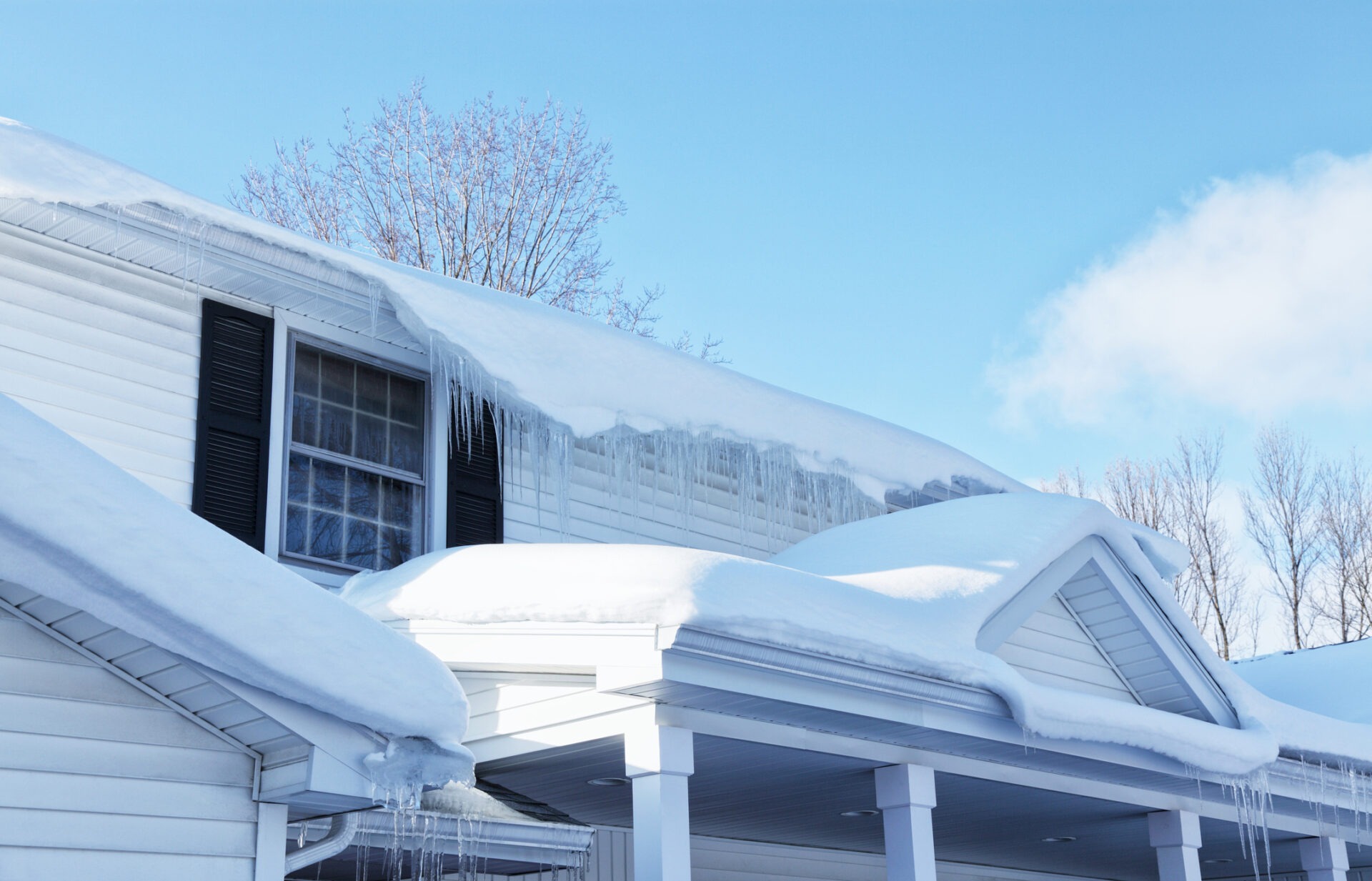 A house with a snow-covered roof and icicles. Clear blue sky and bare trees in the background, creating a winter scene.