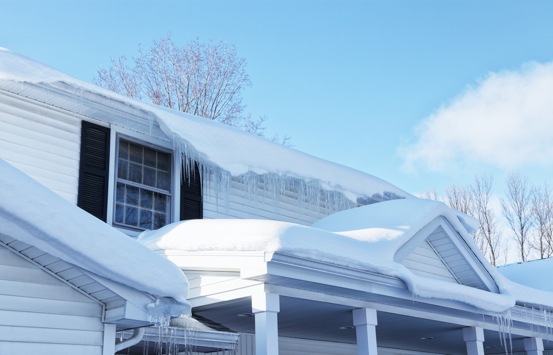 A house with a snow-covered roof and icicles. Clear blue sky and bare trees in the background, creating a winter scene.