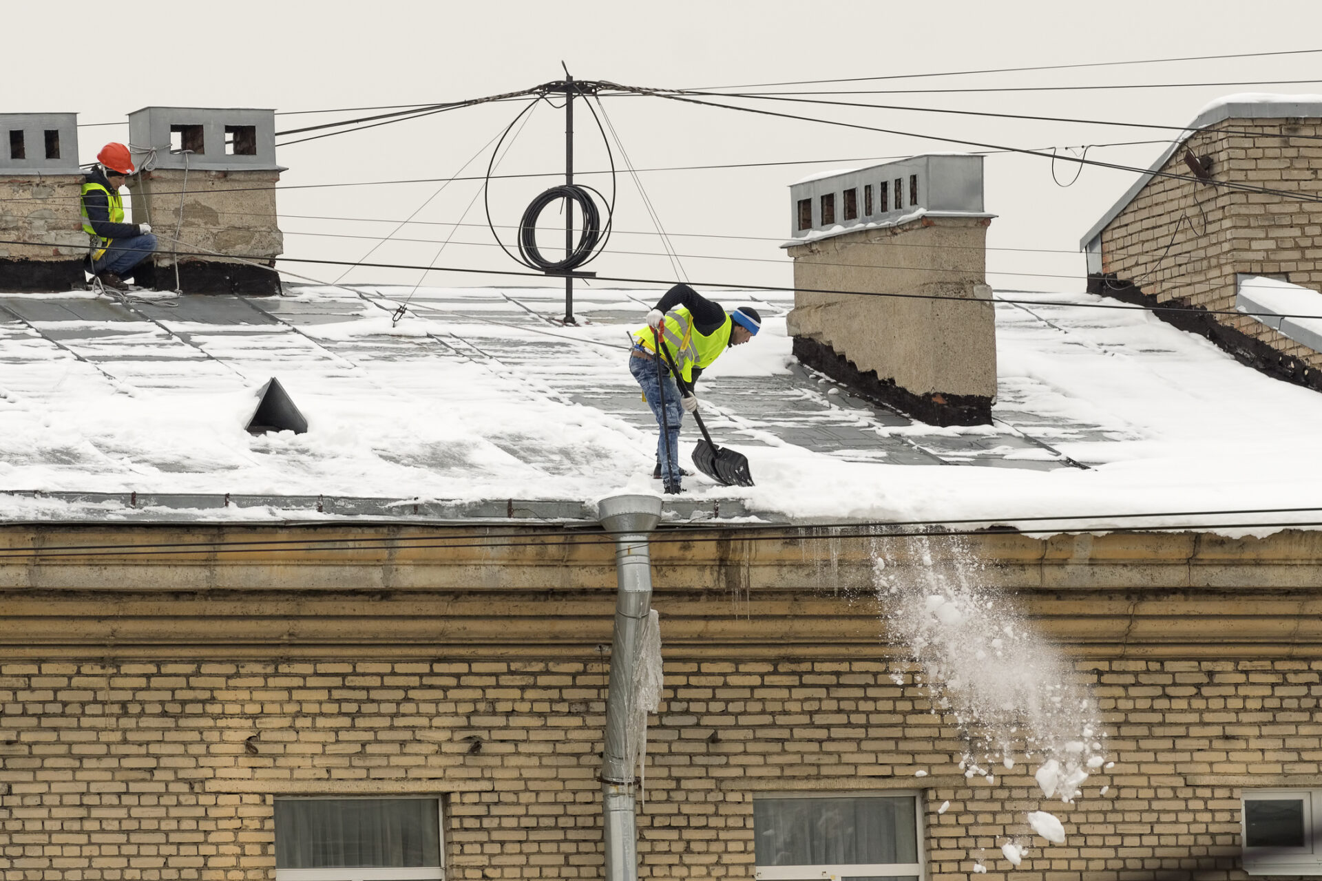 Two persons wearing safety gear clear snow from a building's rooftop, surrounded by brick and concrete chimneys, with snow falling to the ground.