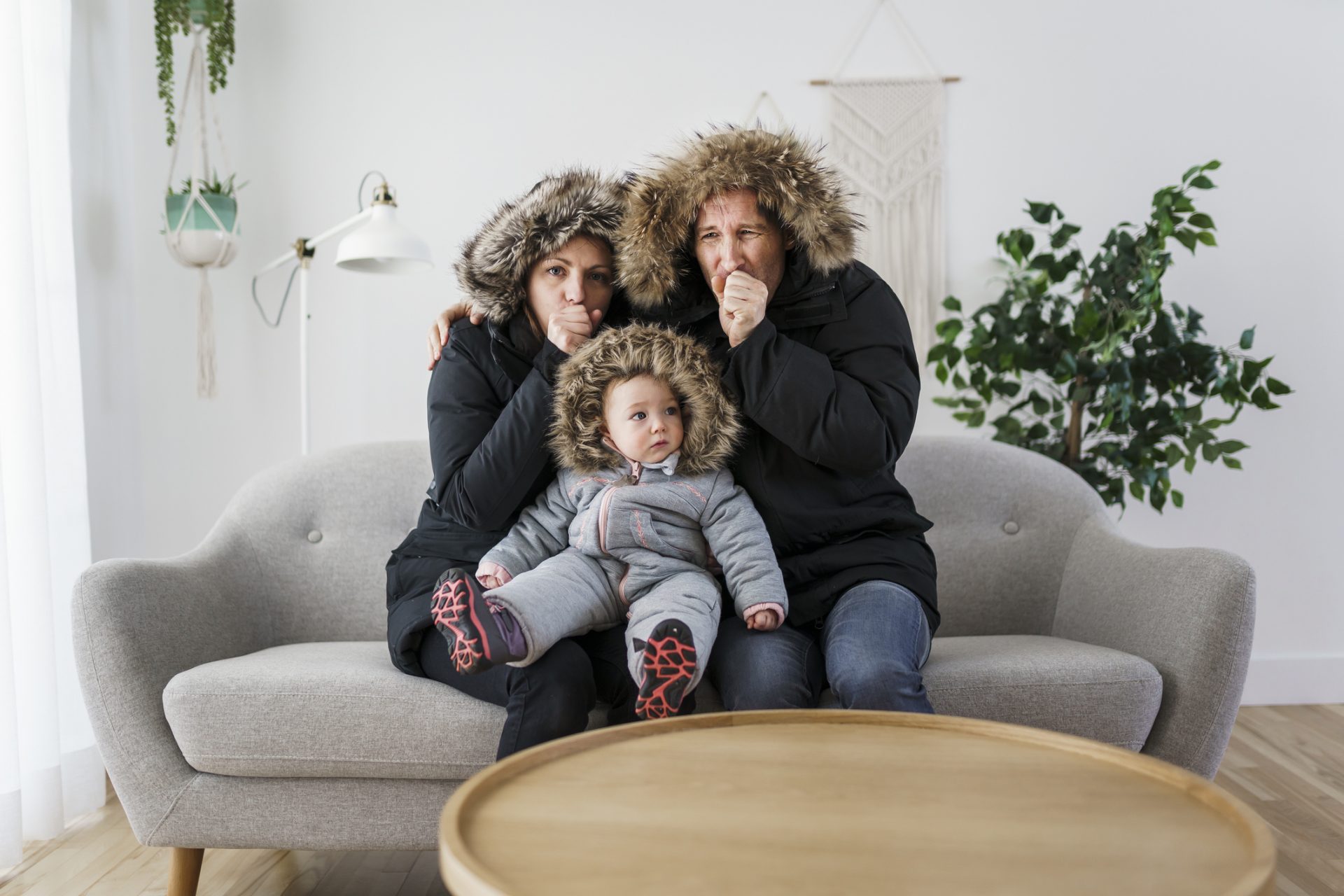 A family huddles together on a couch indoors, wearing winter jackets, blowing on their hands to stay warm.