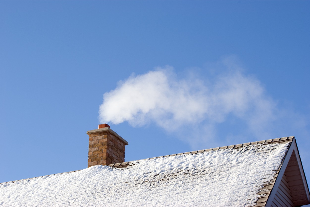 White smoke comes out of a house's chimney on a winter day. The roof is dusted in an even light covering of snow.